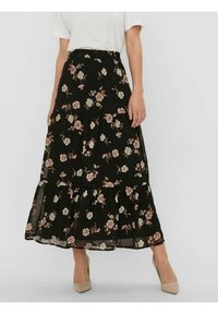 Vrouw in een zwarte bloemenlange midi-jurk met een elastische taille, witte T-shirt met korte mouwen en beige puntige hakken, staande tegen een witte muur.