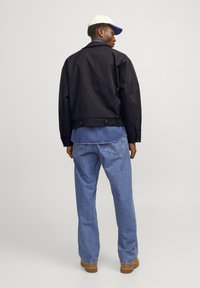 Giacca bomber nera, camicia in denim blu sotto, jeans azzurri chiari e stivali marroni, in piedi contro uno sfondo neutro.