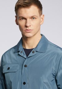 Niebieska, lekka kurtka o gładkiej fakturze. Wyposażona w zapięcie na guzik, dużą kieszeń na piersi oraz kołnierz.