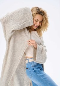 Licht beige opengewerkte cardigan met brede mouwen, gedragen over een aansluitende crèmekleurige top en blauwe jeans. Getextureerde stof met een gezellige uitstraling.