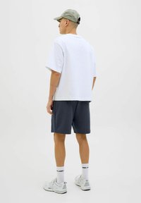 Bijela oversized majica, tamnosive kratke hlače, siva kapa, bijeli atletski tenisice i bijele čarape s crnim tekstom. Običan, ležeran outfit.