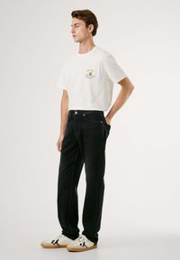 Camiseta de algodón blanca con gráfico frontal, combinada con jeans de mezclilla negros. El modelo lleva zapatillas beige y negras. Fondo neutro.