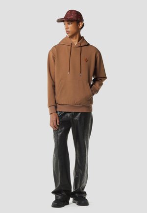 Young man in a brown hoodie, black leather pants, and a patterned maroon cap looking to the side with one hand in his pocket.