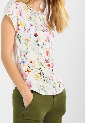 Woman wearing a white short-sleeve top with a colorful floral pattern and green pants, hands casually in pockets.