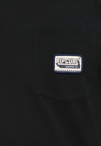 Crna pamučna majica s lijevim džepom. Bijela oznaka sa plavim rubom prikazuje "RIPCURL SURFING CO." izvezeno velikim slovima.