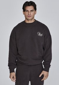 Felpa oversized marrone scuro in tessuto morbido e strutturato. Presenta un colletto a girocollo e un piccolo logo sulla parte sinistra del petto.