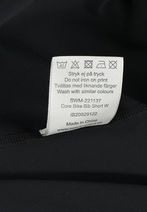 Black bike bib shorts with a care label featuring washing instructions, fabric information, and product code stitched on the inside.