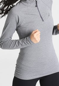 Graues, langärmeliges Sportoberteil mit einem Viertel-Reißverschluss, aus elastischem Material, das einen dezenten Glanz aufweist und ein Logo auf der Brust hat.