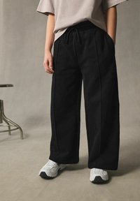 Schwarze weit geschnittene Hose aus weichem Material, mit einem elastischen Bund und Kordelzug sowie seitlichen Taschen, kombiniert mit weißen Sneakers.