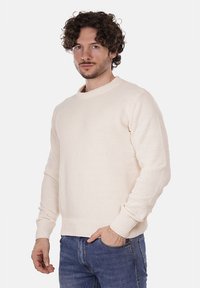 Maglione lavorato a maglia color crema con scollo rotondo e polsini a coste, abbinato a jeans in denim blu. Il tessuto ha una finitura testurizzata.