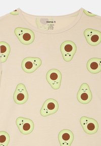 Φωτεινό μπεζ t-shirt με επαναλαμβανόμενο σχέδιο γραφικών αβοκάντο. Διαθέτει πράσινα α�βοκάντο με διάφορες εκφράσεις προσώπου σε απαλό ύφασμα.