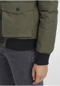 Olive green puffer jacket with a front button closure, ribbed black cuffs, and a patch pocket. Gray denim jeans partially visible.