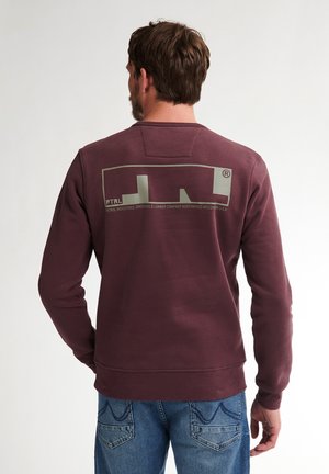 Man wearing burgundy sweatshirt with abstract logo and text on back, paired with blue jeans, standing against a plain light background.