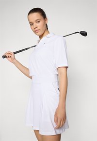 Hvid golf polo med korte ærmer, slim fit og krave, kombineret med en matchende plisseret nederdel. Modellen holder en golfkølle.