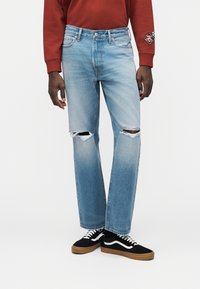 Φωτεινό γαλάζιο τζιν με άνετη εφαρμογή, με σκισίματα στα γόνατα και ξεθωριασμένη όψη, σε συνδυασμό με μαύρα αθλητικά παπούτσια.