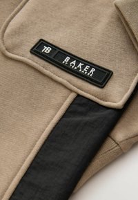Beżowa i czarna tkanina z czarną prostokątną metką z napisem "TB BAKER BY TED BAKER" przyszytą do złożonego odzieży.