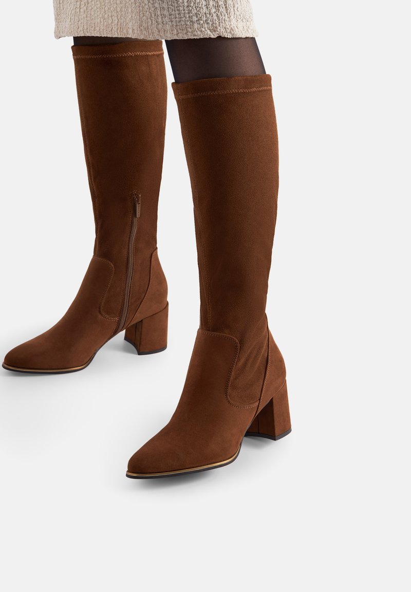 Botas marrones de ante hasta la rodilla con tacones de bloque, cremalleras laterales, usadas con medias negras y una falda beige texturizada.