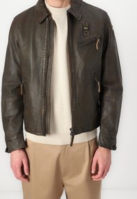 Veste en cuir brun foncé avec un col, fermeture éclair à l'avant et poches latérales. Présente une finition texturée et un ourlet côtelé crème en contraste.