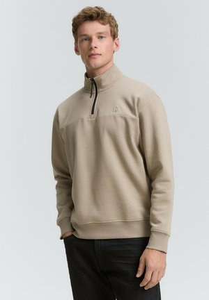 Beige színű, negyed cipzáros pulóver, puha anyagból, texturált panellel a mellkason és egy kis logóval a bal oldalon. Fekete cipzáros részlet.