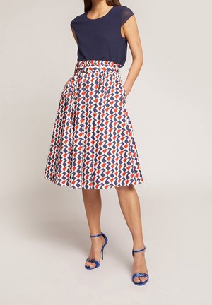 Kvinde iført en marineblå top, knælange nederdel med rødt, hvidt og blåt geometrisk mønster og blå hæle, stående med hænderne i lommerne.