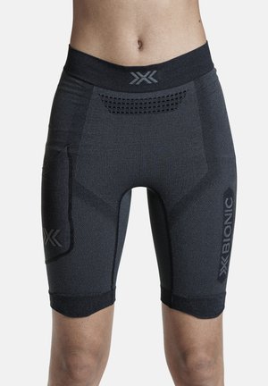 Close-up van een persoon die zwarte compressiebroekjes tot aan de knie draagt met ventilatiedetails en merkslogo’s op de tailleband en dij.