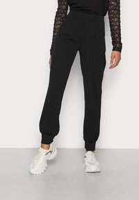 Czarne spodnie dresowe z elastycznymi mankietami przy kostkach, o luźnym kroju i płaskim przodzie. Noszone z jasnymi sportowymi sneakersami.