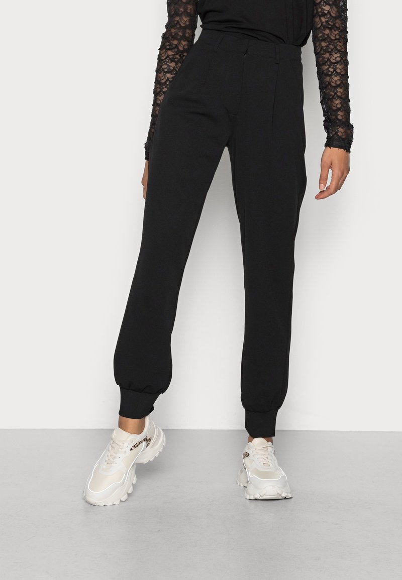 Czarne spodnie dresowe z elastycznymi mankietami przy kostkach, o luźnym kroju i płaskim przodzie. Noszone z jasnymi sportowymi sneakersami.