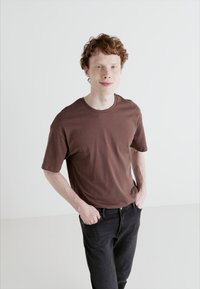 Brown cotton t-shirt with short sleeves, round neckline, and a relaxed fit, paired with dark denim jeans, against a plain white background.