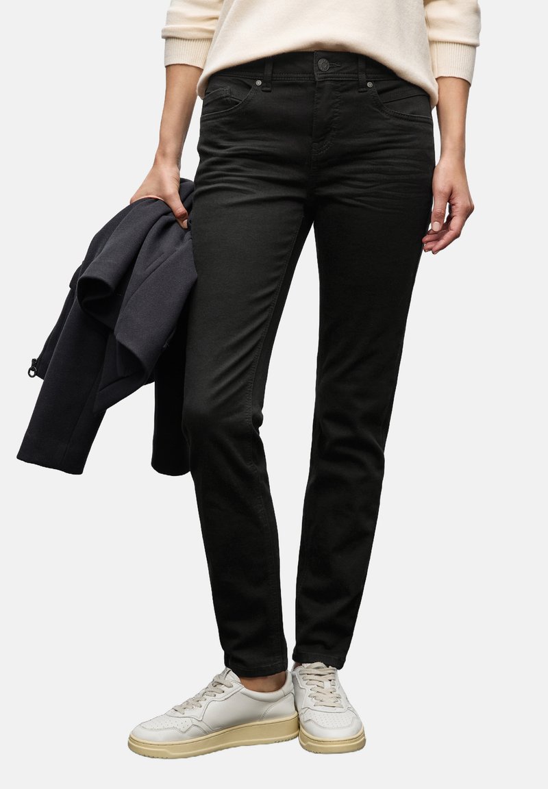 Černé džíny z bavlněné směsi se slim fit střihem, s předními kapsami a pasem na knoflík. Nosič má na sobě světlý svetr a bílé tenisky.