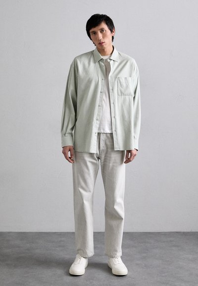 Camicia button-up verde chiaro con taschino sul petto, abbinata a jeans grigi chiari e sneakers bianche. Il tessuto sembra morbido e strutturato.