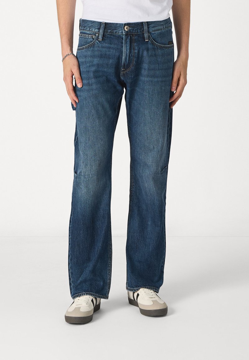 Jeans de color azul oscuro con un diseño de pierna recta, que presentan un clásico patrón de cinco bolsillos, botón de metal y sutiles detalles de desgastado. Combinados con zapatillas blancas.