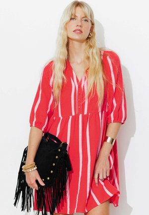 Femme blonde portant une robe rayée rouge et blanche, tenant un sac à main noir à franges, avec des bijoux en or au poignet et aux oreilles.