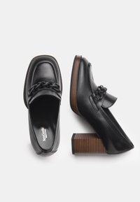 Chaussures à plateforme en cuir noir avec un nez carré, un accent chaîne sur le dessus et un talon bloc en bois rayé. Texture lisse et design épuré.