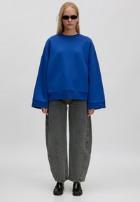 Blaues Oversized-Sweatshirt mit weiten Ärmeln, kombiniert mit hochgeschnittenen, lockeren grauen Jeans. Das Model trägt schwarze Sonnenbrillen und Schuhe.