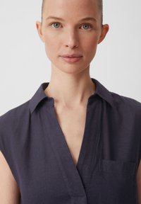 Marineblaue, ärmellose Bluse mit V-Ausschnitt, Kragen und Brusttasche; gefertigt aus strukturiertem Stoff mit Steppdetails.