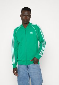 Zaļa zip-up jaka ar baltām trīs svītru akcentiem uz piedurknēm. Ietver augstu apkakli un adidas logo uz krūtīm. Mīksta auduma tekstūra.