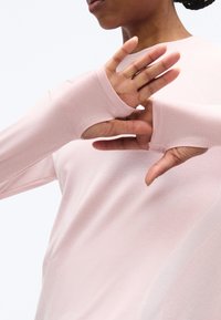 Άτομο με ροζ ανοιχτόχρωμο μακρυμάνικο μπλουζάκι με τρύπες για τους αντίχειρες, τα χέρια σηκωμένα κοντά στο πρόσωπο, παρουσιάζοντας λεπτομερή υφή του υφάσματος και τον τόνο του δέρματος.