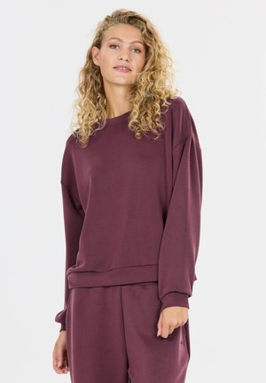 Femme aux cheveux bouclés portant un haut bordeaux ample à manches longues et un pantalon assorti, debout devant un fond clair uni.