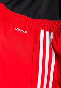 Shorts de sport rouges avec des rayures verticales blanches et une ceinture élastique, avec le mot "AEROREADY" en petites lettres blanches près de la taille.