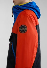 Chaqueta a bloques de color con paneles en rojo, azul y negro. Fabricada en material ligero, cuenta con un parche de logo y una capucha ajustable con cordón.