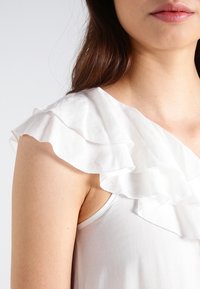 Top blanco sin mangas con detalle de volantes en el hombro. Textura suave, tejido blando, diseño ajustado y escote amplio.