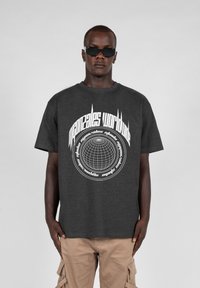 Dunkelgraues Baumwoll-T-Shirt mit kurzen Ärmeln, das in der Mitte einen weißen grafischen Text und ein Globusdesign zeigt. Kombination mit beigen Cargo-Shorts.