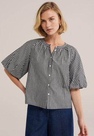 Femme blonde portant un chemisier à manches courtes rayé vertical noir et blanc avec des boutons, associé à un pantalon bleu foncé, debout la main sur la hanche.