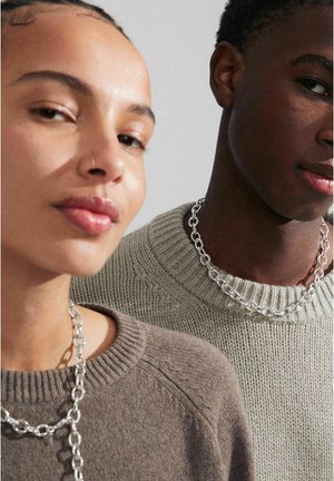 Two models wear textured knit sweaters—one brown, one light gray. Both sport chunky silver chain necklaces, showing their necklines.