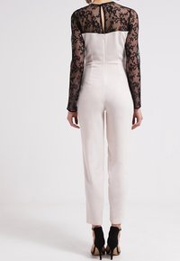 Lace long-sleeve jumpsuit in cream with a fitted silhouette, featuring a sheer black lace top and tapered legs, paired with black heels.