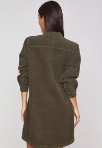 Abito chemisier in cordura verde oliva con una vestibilità rilassata, maniche lunghe arrotolate e un dettaglio di yoke sul retro. Tessuto testurizzato con un motivo a righe verticali.