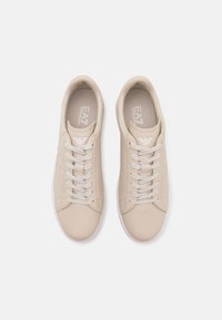 Béžové tenisky na šněrování s hladkým syntetickým svrškem, bílou podrážkou a raženým logem. Texturované detaily po stranách zlepšují design.
