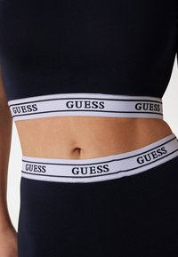 Crni pamučni crop top s bijelom brendirano trakom na kojoj piše "GUESS" crnim slovima. Glatka tekstura, pripijen dizajn s izrezom u sredini trbuha.