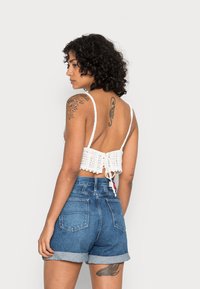 Top cropped de crochê branco com laços ajustáveis nas costas, combinado com calções de ganga de cintura alta azuis com barras dobradas.