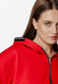 Czerwona bluza z kapturem na zamek, o gładkiej fakturze, z czarną metką z logo przy szyi i usztywnionym kapturem.